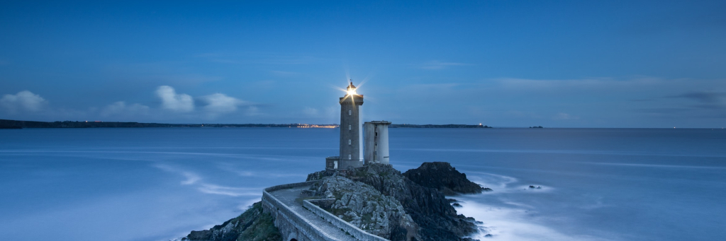 Quay wall leading to Lighthouse at blue lit dawn with smooth ocean and distant shore city.